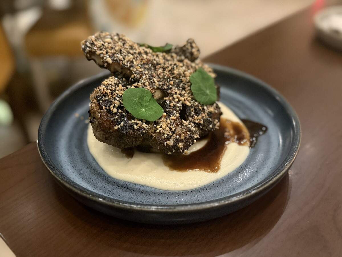 A close-up of a sophisticated dish served in a dark blue bowl on a polished wooden table. The meal features a piece of cauliflower heavily crusted in a sesame and nut mixture, resting atop a smooth, pale purée. A dark, rich sauce is drizzled around the base, and the dish is garnished with two bright green round leaves.