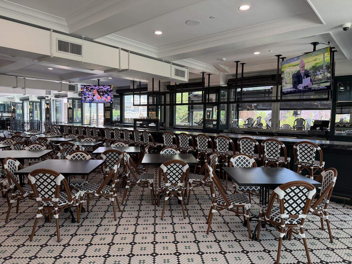 A view of a long, dark marble bar with several wall-mounted televisions showing news and sports. The foreground is filled with black-topped tables and bistro chairs with a black-and-white geometric weave, set against a detailed mosaic tile floor.