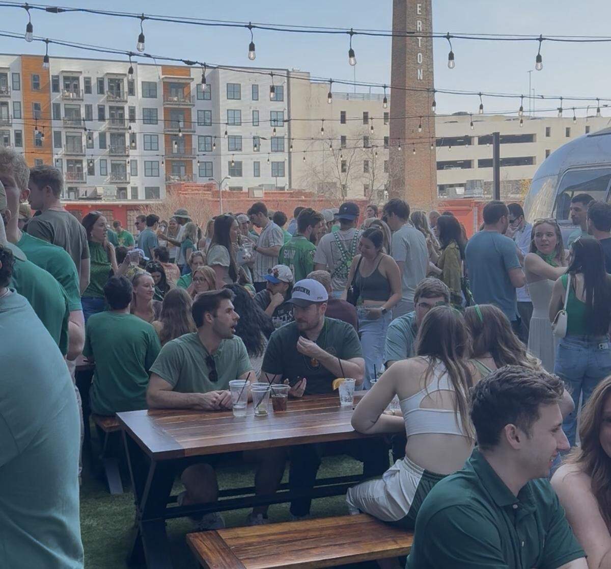 A crowded, outdoor patio area filled with people, most of whom are wearing green shirts. The scene is bright and sunny, with string lights overhead and a large brick chimney in the background. In the foreground, several people are seated at wooden picnic tables talking and drinking. In the background, there is a large apartment building and a parking garage. The overall atmosphere is lively and social.
