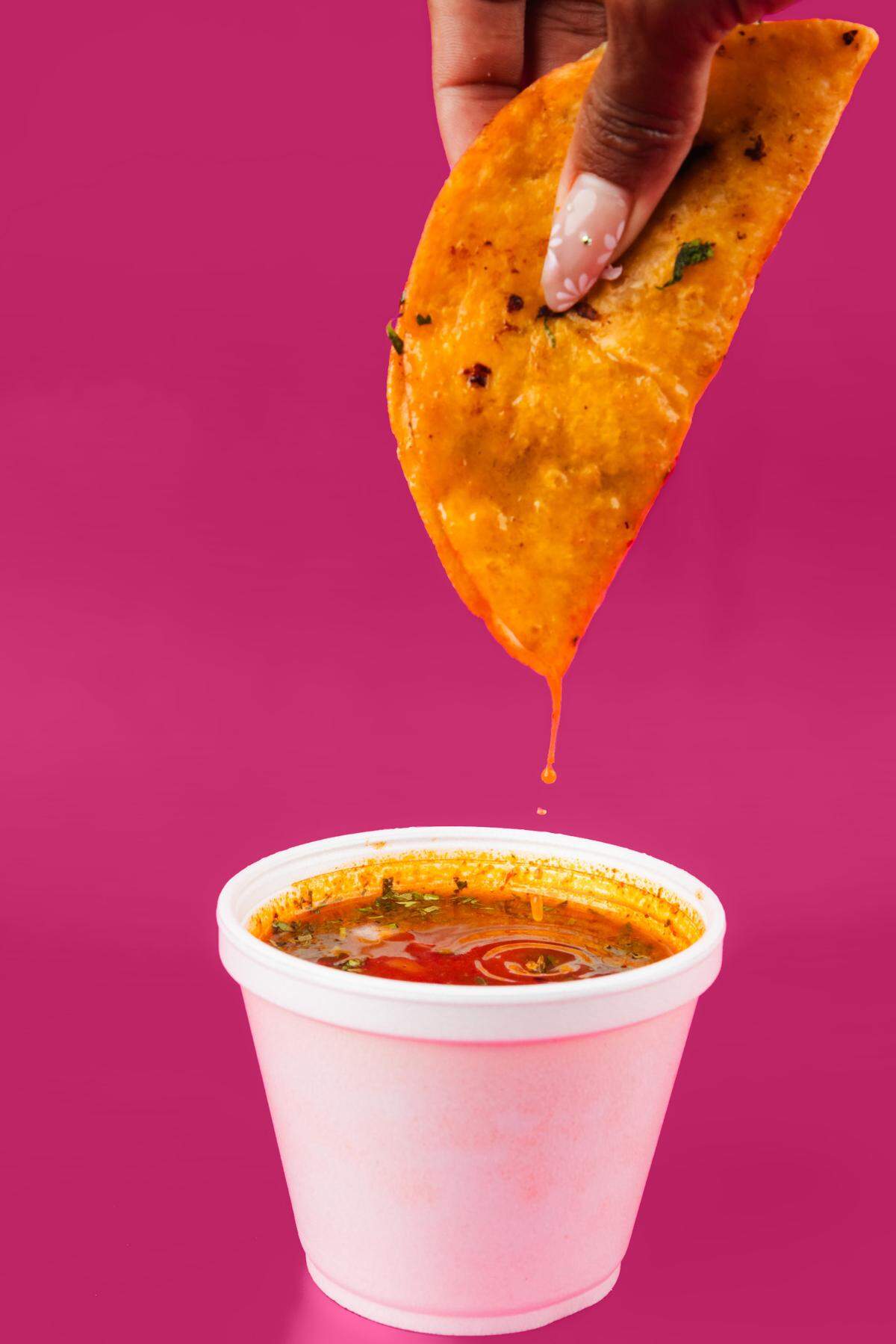 Alt Text: A close-up shot against a solid bright pink background. A hand with manicured nails holds a golden-brown, oil-dipped birria taco, which is dripping juice into a white foam cup filled with rich, red consomé broth topped with fresh cilantro.