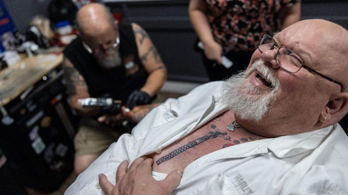 Paul Elkins laughs as he shows off a zipper tattoo over an open heart surgery scare that Jeff Brown, left, tattooed for him at Tattoo Bill’s in 2023.