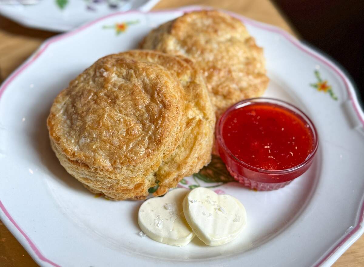 Two large, golden-brown flaky buttermilk biscuits served on a vintage white plate with a pink rim. The plate also holds a small glass bowl of vibrant red jam and two heart-shaped pats of butter topped with flaky sea salt.