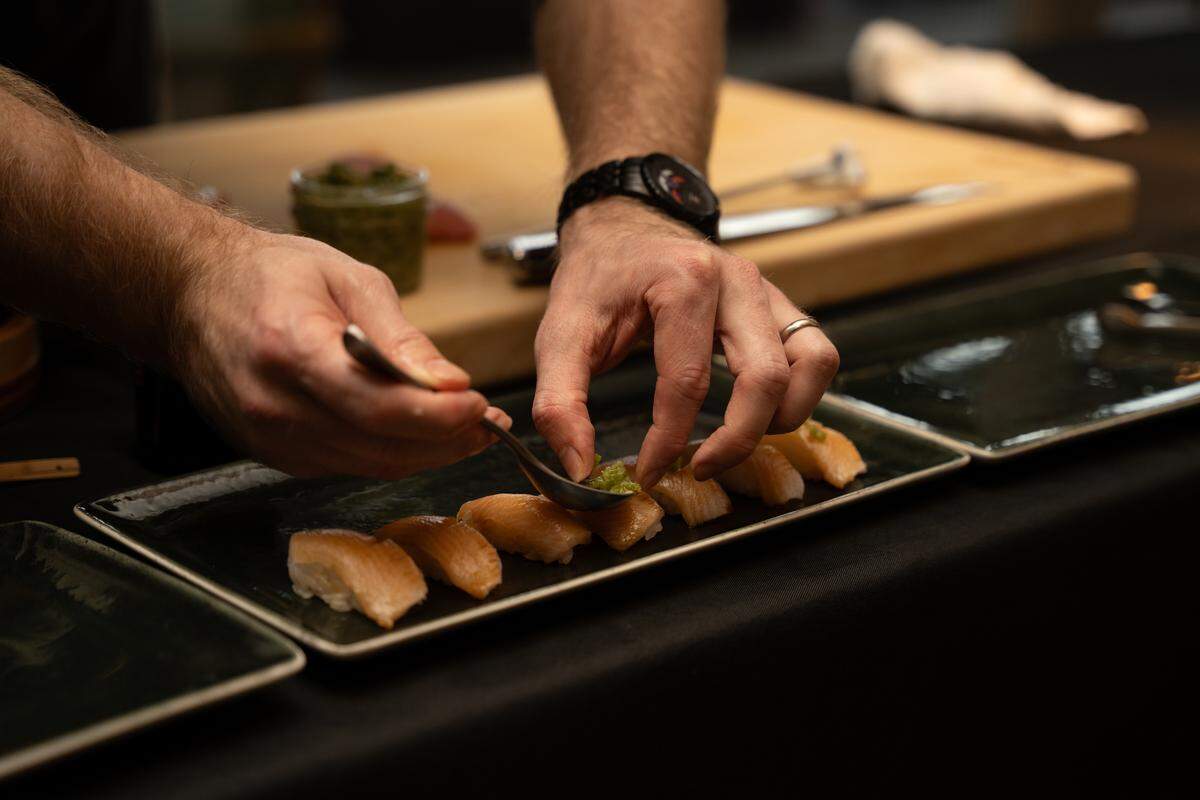A close-up of a chef’s hands using a small spoon to carefully place a green garnish onto several pieces of nigiri lined up on a dark rectangular plate.