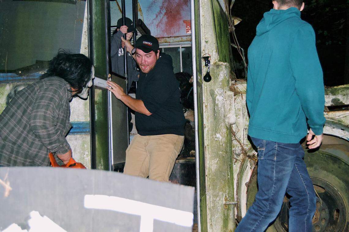 In a dark, outdoor setting, a person in a plaid shirt and a white mask wields an orange chainsaw while approaching the doorway of an old, dilapidated bus. A person in a black hoodie and baseball cap is exiting the bus with a look of alarm, while another person in a teal hoodie walks away in the foreground.