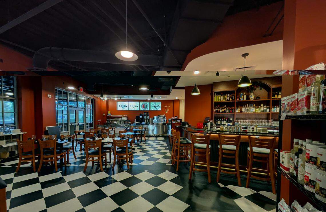 The interior of the “Osteria” dining and bar area. The room features terracotta-colored walls, a black and white checkered floor, and dark industrial ceilings. To the right is a wood-topped bar with several wooden stools, while the left side is filled with wooden dining tables and chairs.