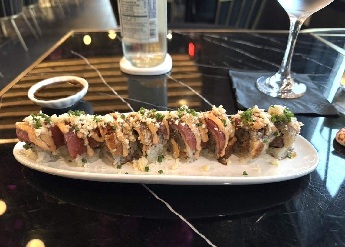 A sushi roll topped with spicy mayo, crispy tempura flakes, and chopped chives is served on a white plate with a small dish of soy sauce. A glass of water and a bottle sit on a glossy black marble table in the background.