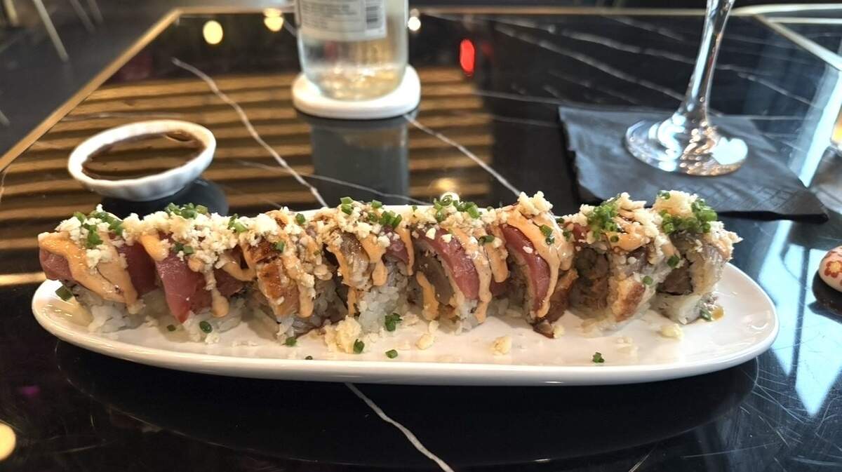 A sushi roll topped with spicy mayo, crispy tempura flakes, and chopped chives is served on a white plate with a small dish of soy sauce. A glass of water and a bottle sit on a glossy black marble table in the background.