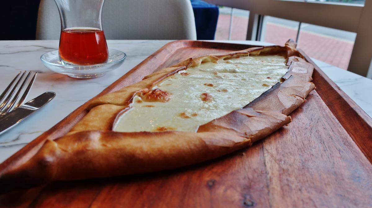 A Turkish cheese pide, a long, boat-shaped flatbread filled with melted white cheese, is served on a wooden board next to a glass of Turkish tea.