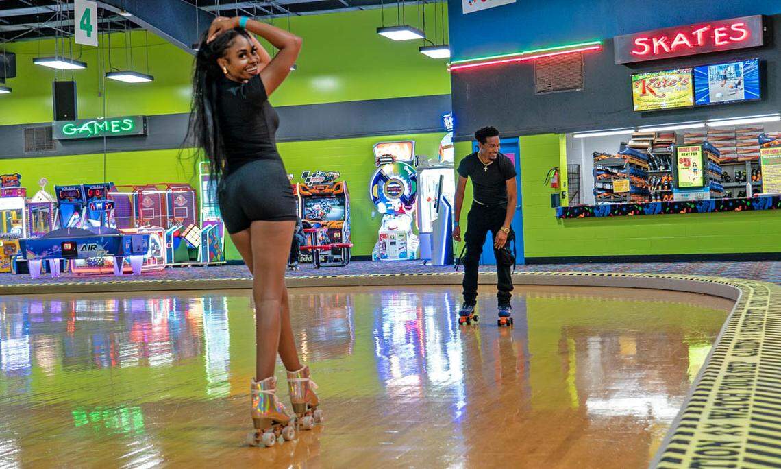 A lively, wide shot of an indoor roller skating rink. In the foreground, a woman wearing a black outfit and roller skates poses for the camera, smiling with her hands on her head and hip. In the midground, a man in a black outfit roller skates on the polished wooden floor, smiling as he looks towards her. The background features a vibrant green wall, an arcade area with a green neon “GAMES” sign on the left, and a skate rental counter with a red neon “SKATES” sign for “Kate’s” on the right.