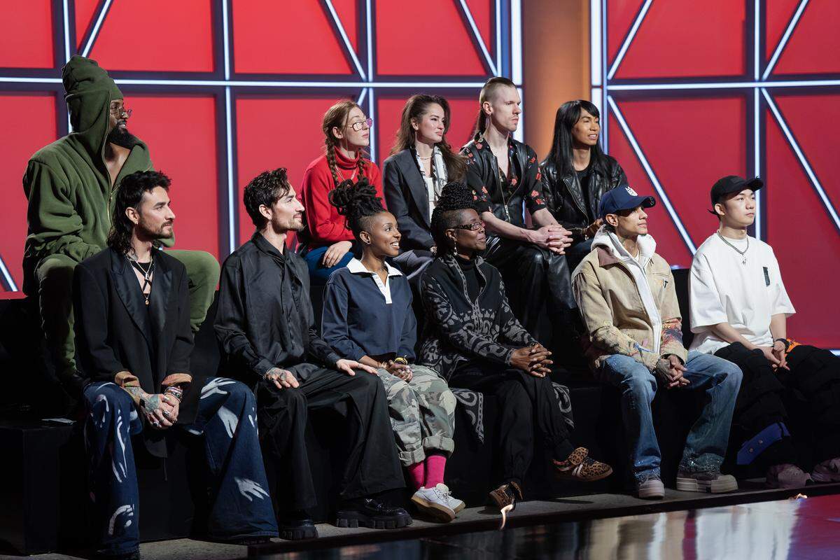 Eleven people sit on two levels facing something that is off camera. Two are wearing hats, one a hood and they have fashionable clothes on, mostly in black but one person is wearing a red turtleneck. Another is wearing a dark green robe, one a white t-shirt and another a tan jacket. The wall behind is bright red with black and white lines for decor.