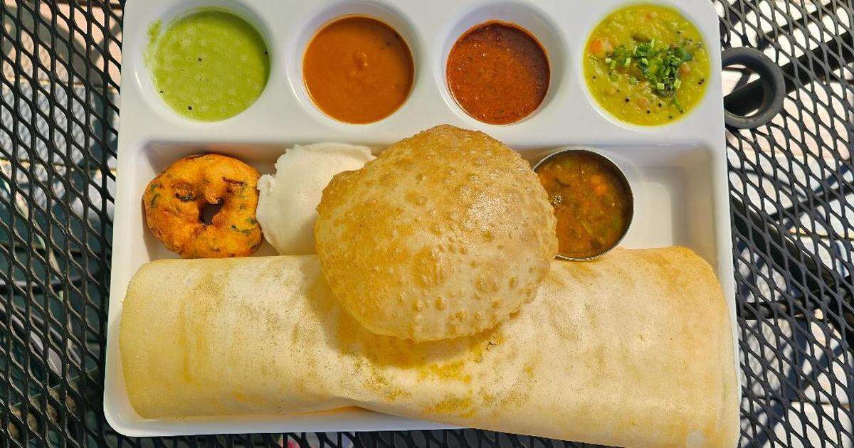 A rectangular white platter filled with South Indian breakfast items (tiffin). It includes a long rolled-up dosa, a puffed poori and small idli resting on the dosa, a vada on the left, and several small bowls of colorful accompaniments (green chutney, red chutneys, and sambar). The platter is sitting on a black, wire-mesh outdoor table.