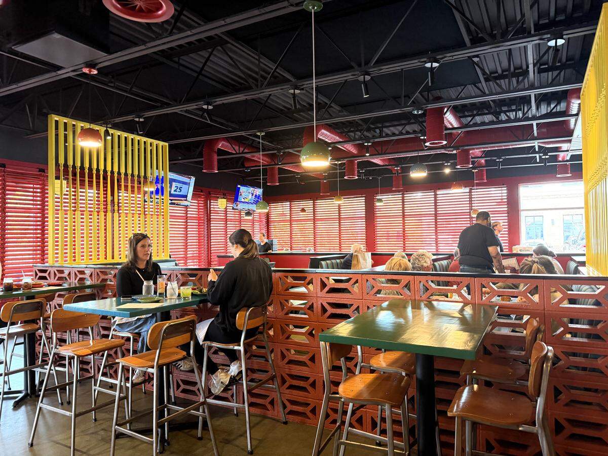 The spacious dining room of Chili Willis showing a mix of high-top tables and booths. The space features industrial black ceilings with red ductwork, red window shutters, and a warm, eclectic color palette of green, yellow, and orange. Several patrons are seated and dining throughout the room.