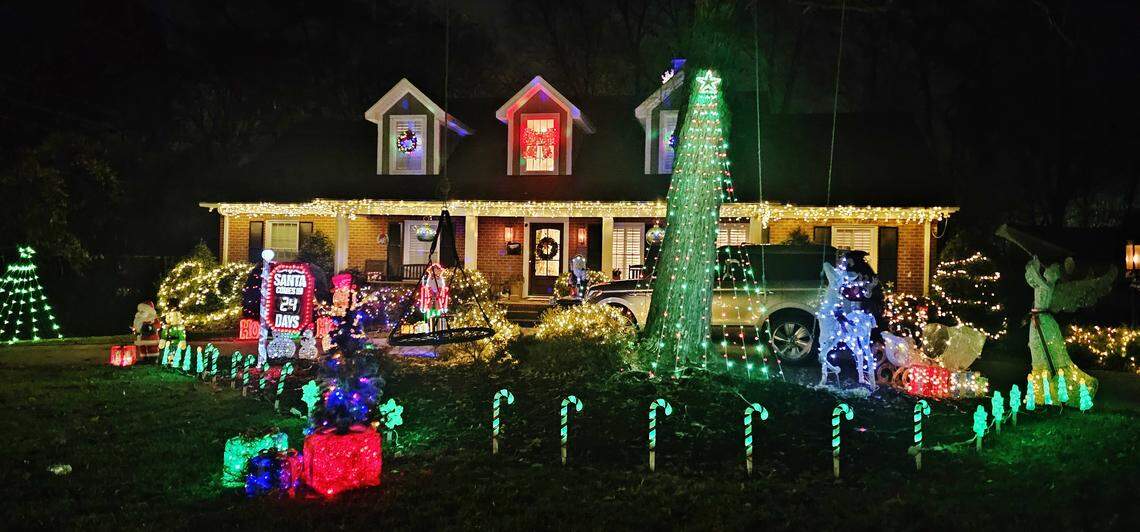 A two-story home brightly decorated for Christmas at night. The lawn features a large display with a glowing green archway tree, a lit-up sign counting down the days until Christmas, several candy cane lights lining the sidewalk, and numerous brightly colored figures and wrapped light bushes.