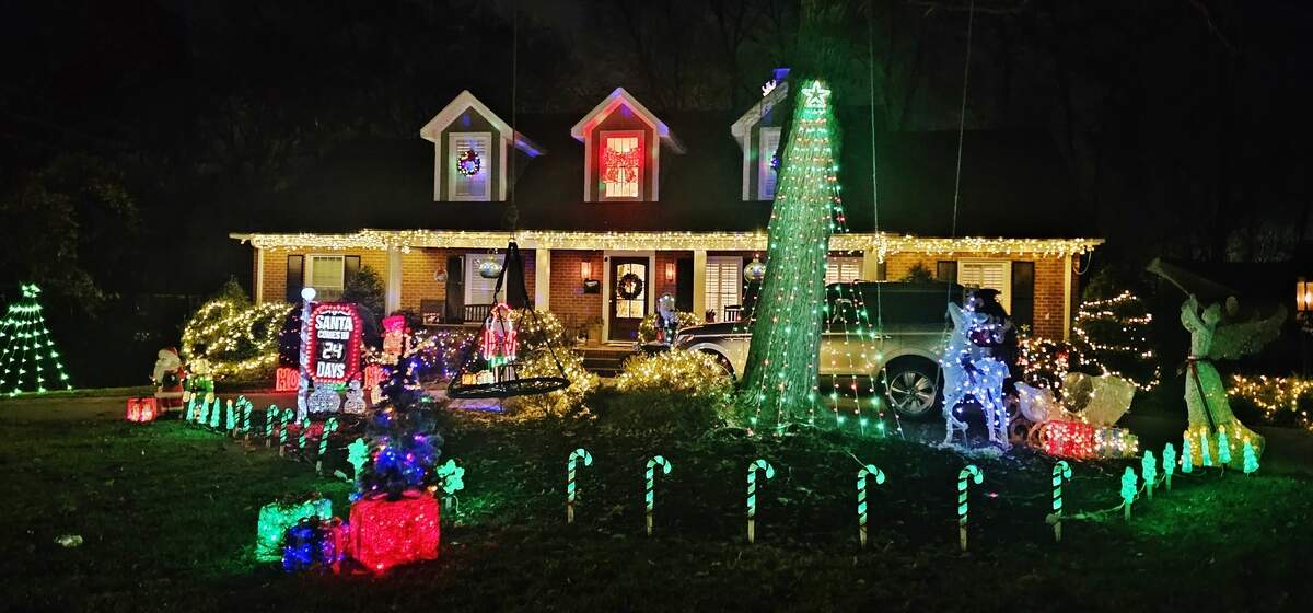 A two-story home brightly decorated for Christmas at night. The lawn features a large display with a glowing green archway tree, a lit-up sign counting down the days until Christmas, several candy cane lights lining the sidewalk, and numerous brightly colored figures and wrapped light bushes.