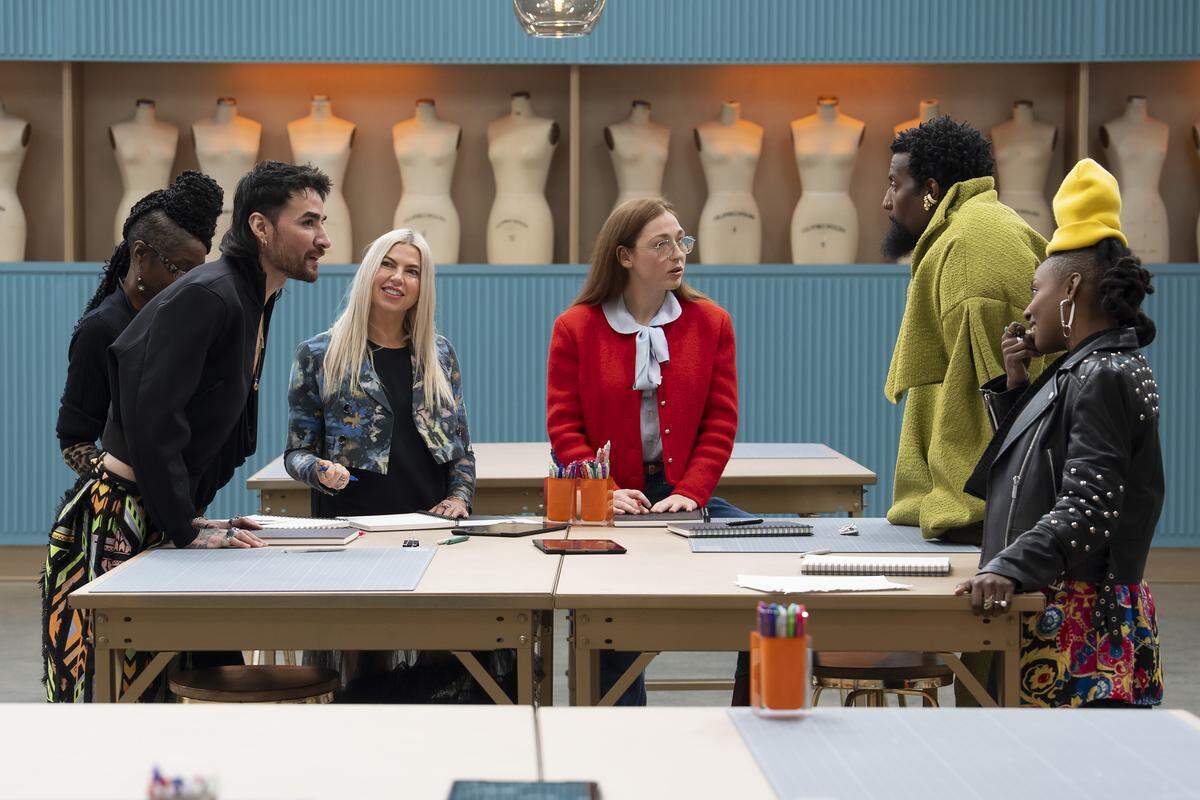 Six adults stand around a folding table wearing fashionable outfits made up of black, green, orange, red, yellow and pink. Some of the people seem to be talking with others listening. A folding table is in the foreground, another is in the background. The wall behind is turquoise with a ribbed texture. A beige cutout in the wall holds 12 empty mannequin busts.