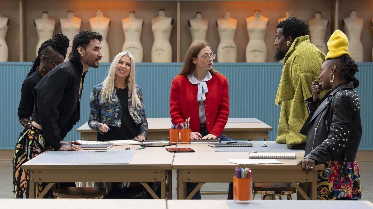 Six adults stand around a folding table wearing fashionable outfits made up of black, green, orange, red, yellow and pink. Some of the people seem to be talking with others listening. A folding table is in the foreground, another is in the background. The wall behind is turquoise with a ribbed texture. A beige cutout in the wall holds 12 empty mannequin busts.