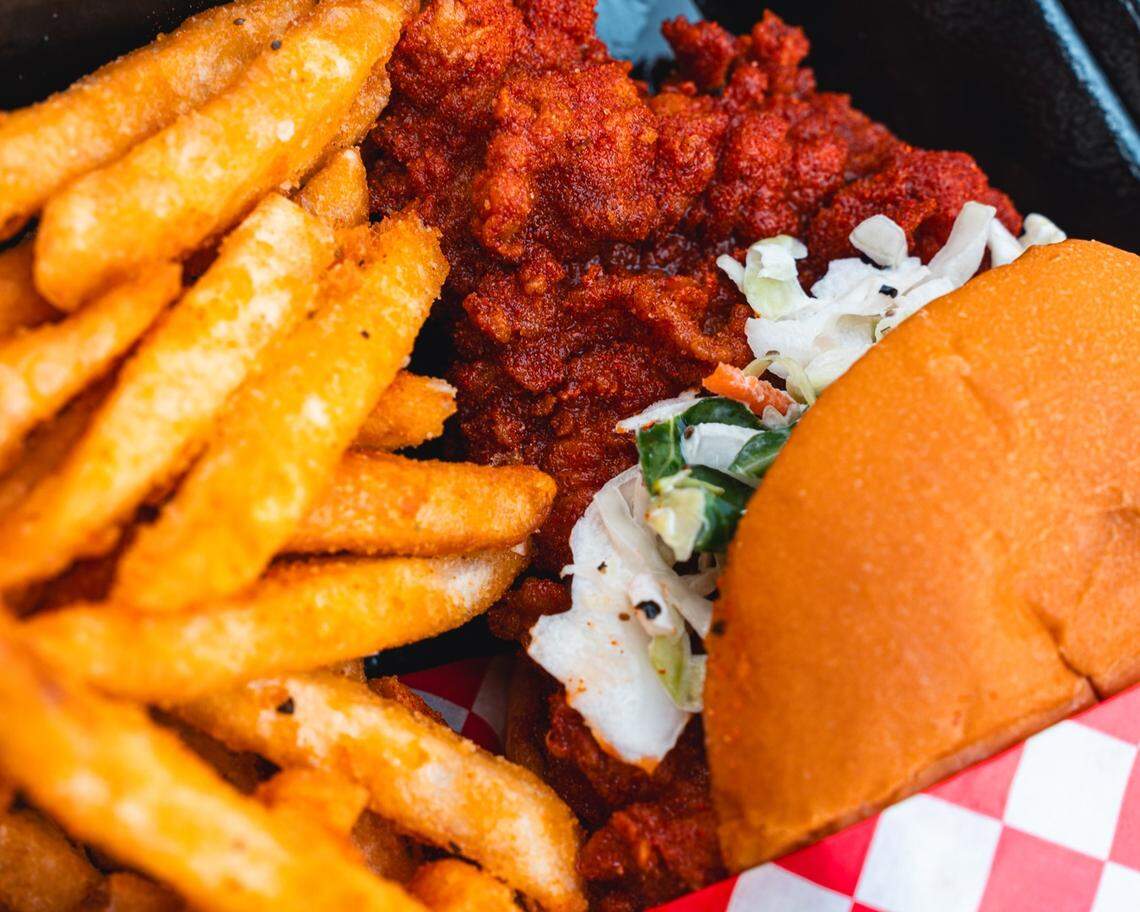 A close-up overhead shot of a spicy Nashville hot chicken sandwich with coleslaw, served next to a generous portion of seasoned french fries in a container lined with red and white checkered paper.