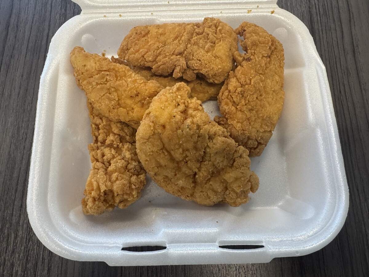 A top-down view of five golden-brown fried chicken tenders served inside an open, square white styrofoam takeout container. The chicken pieces have a light, evenly textured, and crispy breading and are stacked loosely in the center of the container.