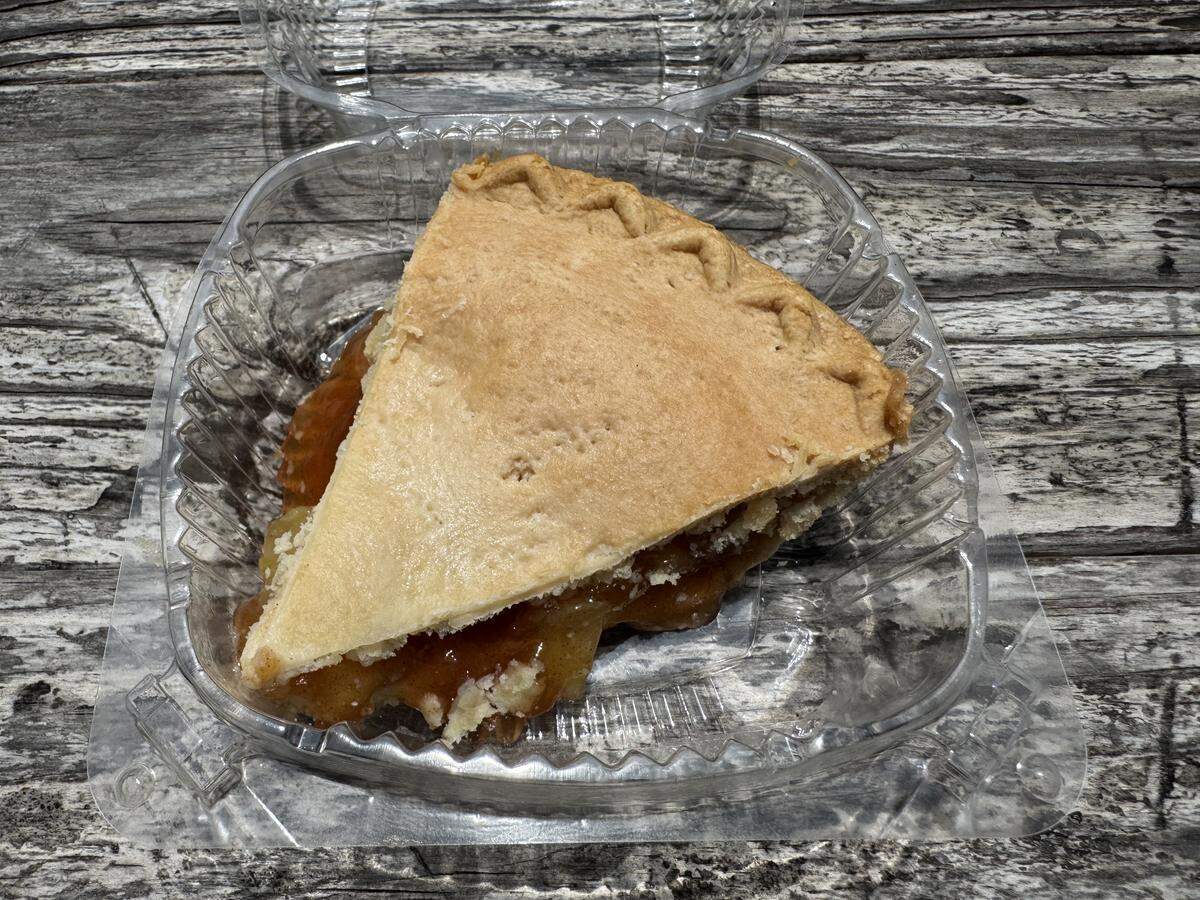 A close-up shot of a single slice of fruit pie with a golden-brown, crimped top crust inside a clear plastic clam-shell takeout container. Thick, glossy fruit filling and pieces of lower crust are visible along the sides. The container sits on a rustic, gray-toned wooden table.