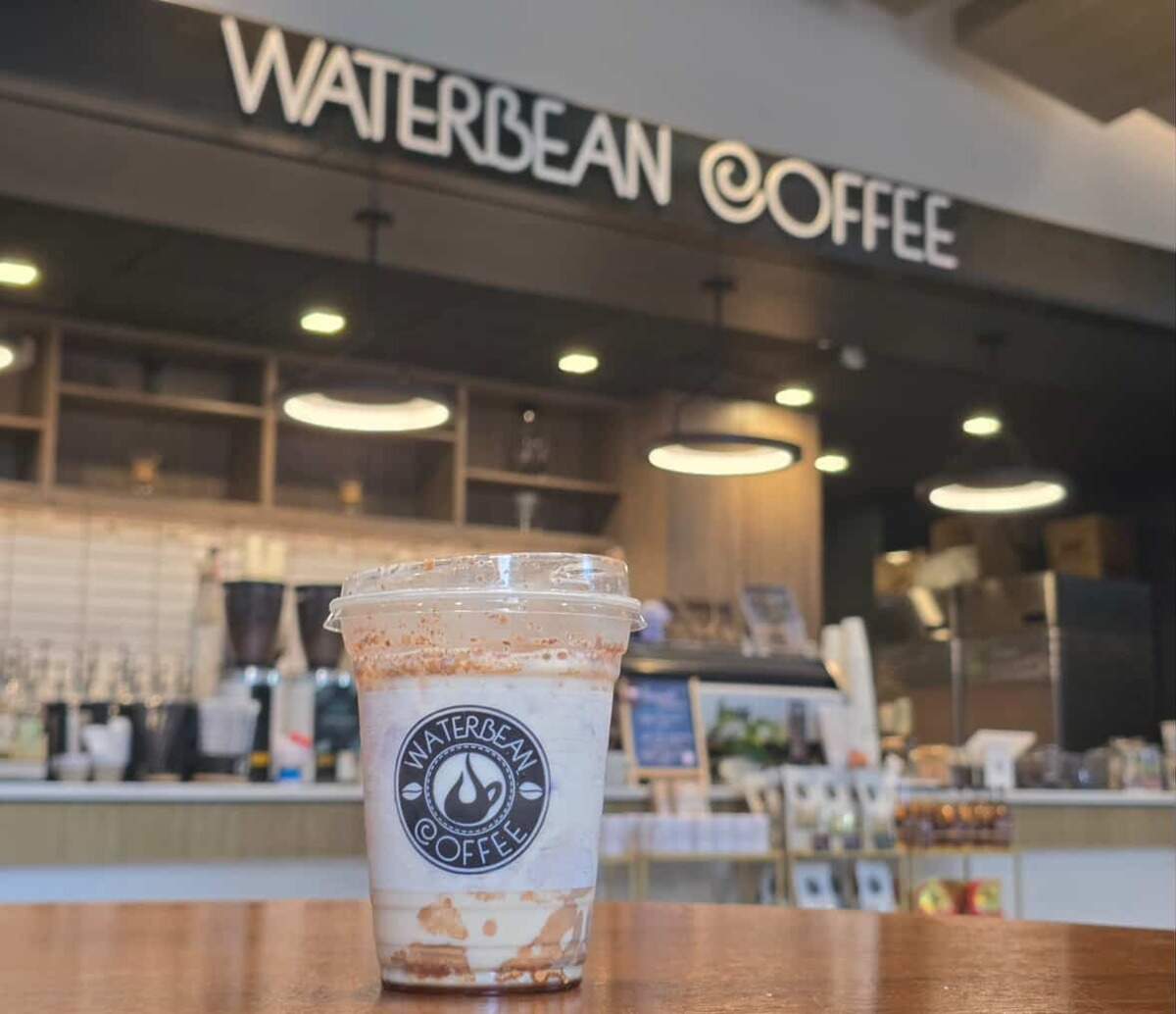 An inviting photo of a specialty iced coffee drink from Waterbean Coffee. The branded cup is featured in the foreground on a wood table, with the bright, modern interior of the coffee shop and its sign creating a warm and welcoming backdrop.