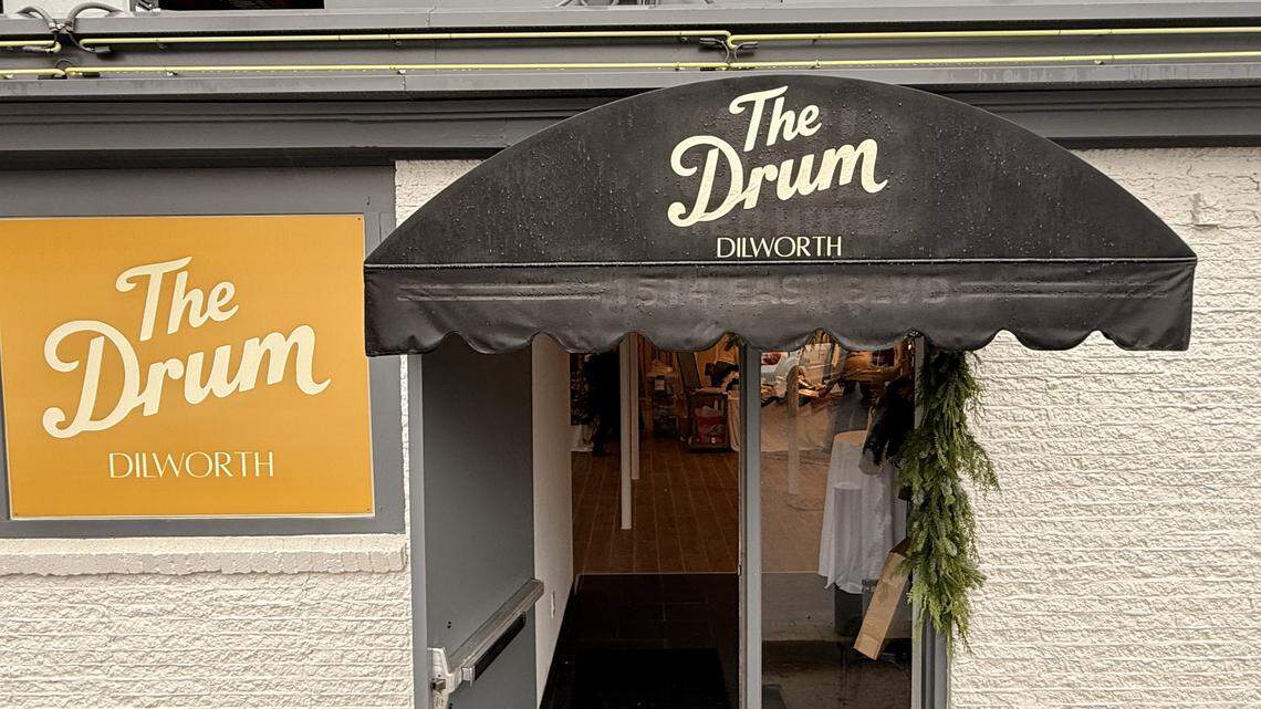 The exterior of a building with white, textured siding. A black, scalloped awning hangs over the entrance door, displaying the text “The Drum DILWORTH” and the address “1514 EAST BLVD” in white script. To the left of the door, an orange square sign also reads “The Drum DILWORTH” in white. A glimpse of the bright interior is visible through the doorway, which is decorated with a strand of greenery.