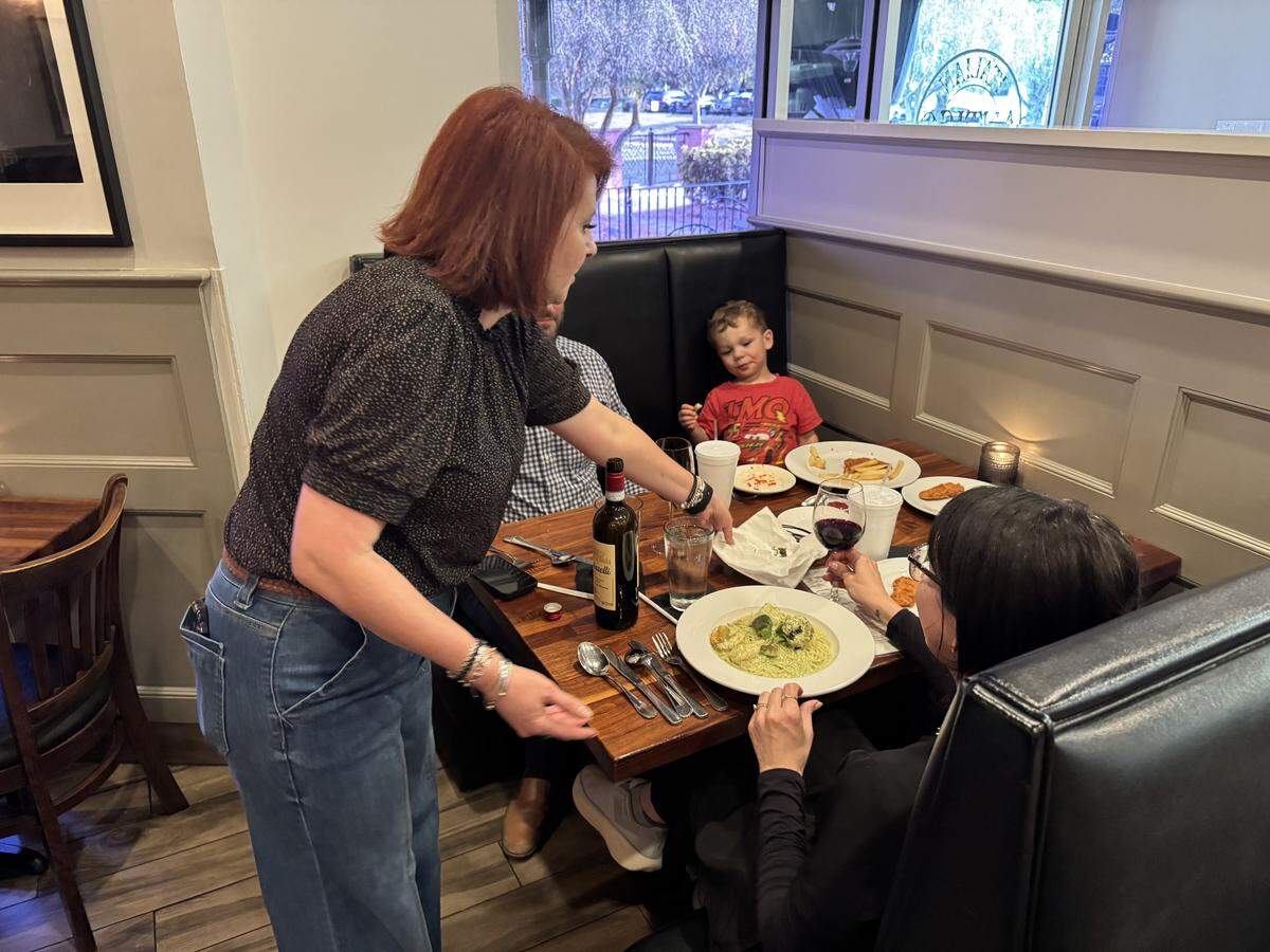 A server with red hair leans over a wooden restaurant table to serve a family seated in a black leather booth. On the table, there is a bottle of wine, a glass of red wine, and a plate of pasta with green pesto sauce. A young child in a red shirt is seated at the far end of the table.