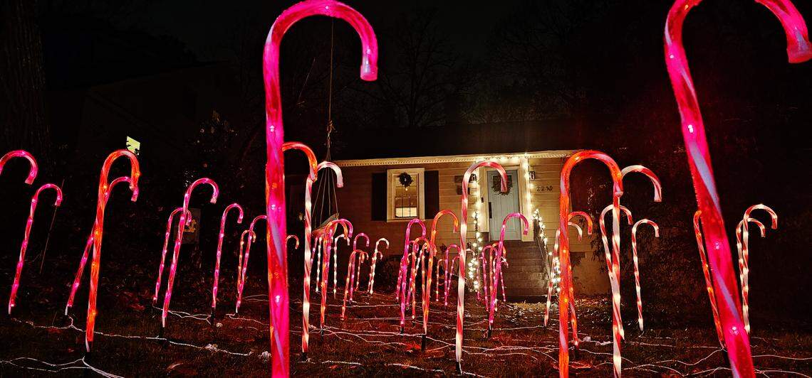 A front yard illuminated by a dense installation of towering, luminous pink and red candy cane decorations. The path of candy canes leads up to the entrance of a small, single-story home with a wreath on the front door, creating a whimsical and surreal holiday scene.