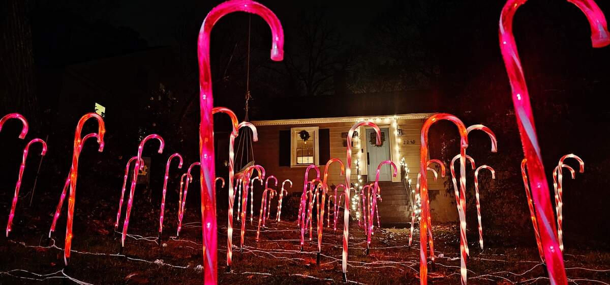 A front yard illuminated by a dense installation of towering, luminous pink and red candy cane decorations. The path of candy canes leads up to the entrance of a small, single-story home with a wreath on the front door, creating a whimsical and surreal holiday scene.
