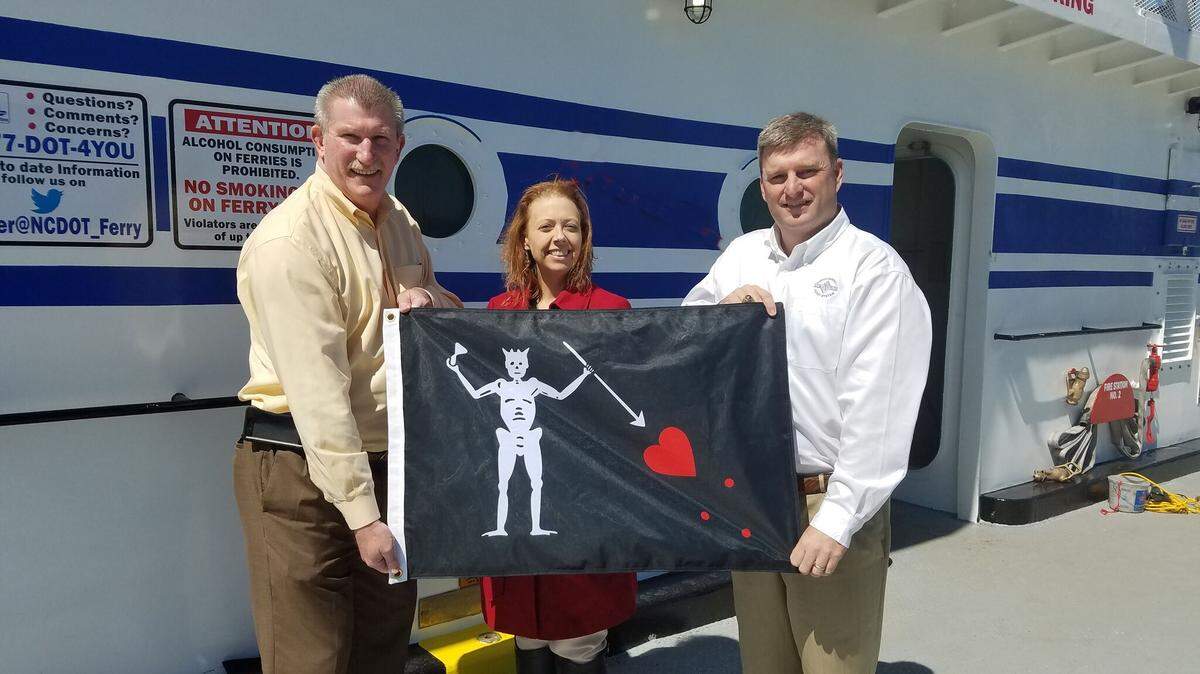 N.C. Ferry Division director Harold Thomas, Blackbeard 300 Committee chair LeRae Umfleet and deputy ferry director Jed Dixon with one of the Blackbeard flags that will fly on North Carolina ferries this year.