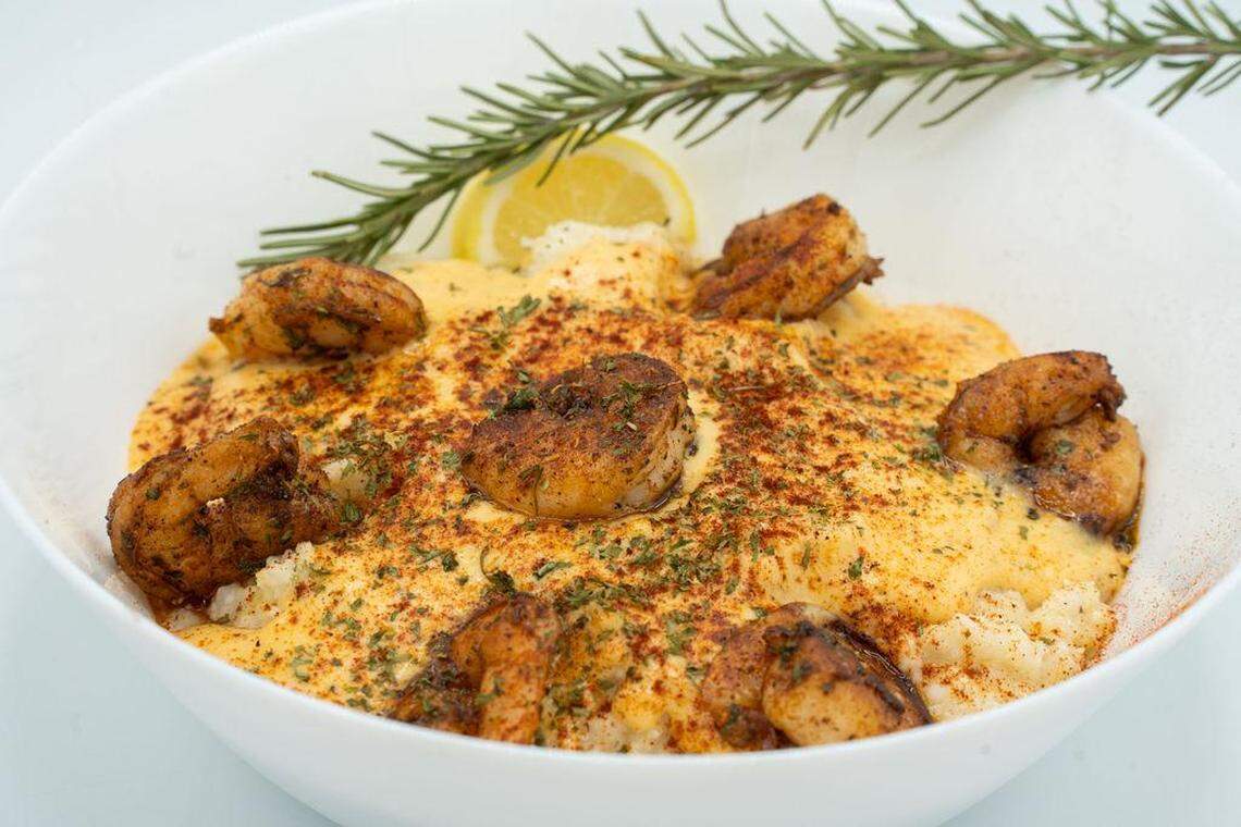 A white bowl filled with creamy yellow grits topped with blackened shrimp and a rich sauce. The dish is finished with a dusting of paprika, fresh herbs, a lemon wedge, and a long sprig of fresh rosemary leaning against the rim.