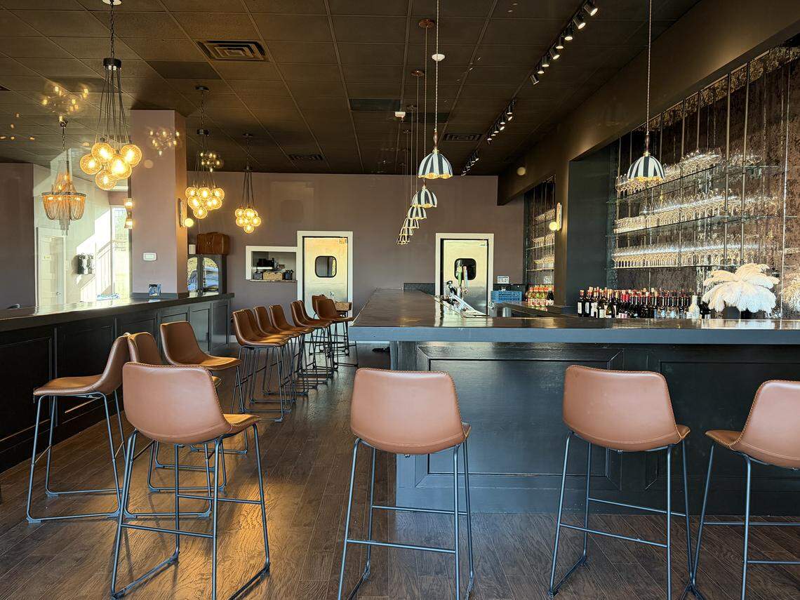 An interior view of a modern wine bar featuring a long, black L-shaped counter. Warm, tan leather barstools with thin black metal legs are lined up along the bar. The back wall is painted a muted plum color and features a large, back-lit glass shelving unit filled with wine glasses. Decorative lighting includes a mix of orb-style chandeliers and black-and-white striped pendant lights. The floor is dark hardwood under a black industrial ceiling.
