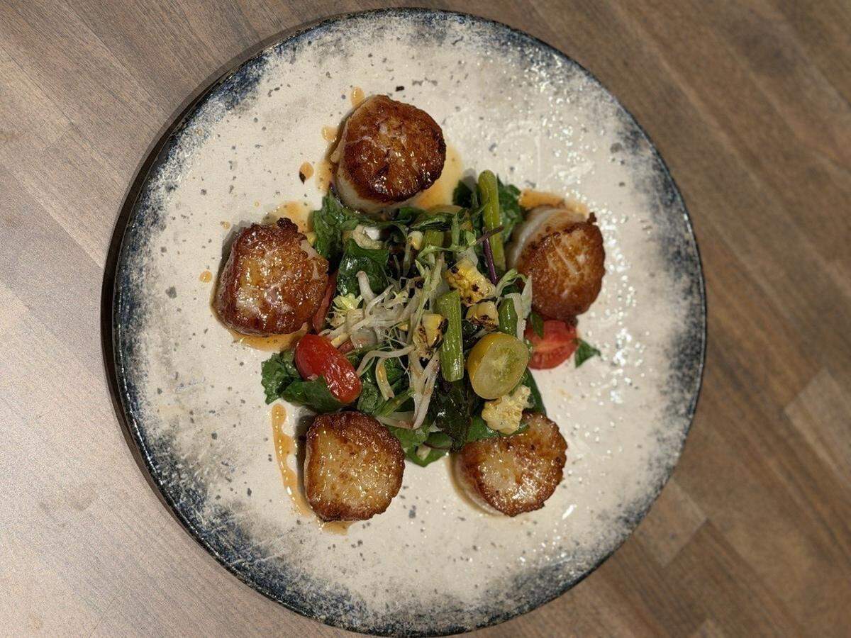 Five seared scallops are artfully arranged in a circle around a central pile of greens and sautéed vegetables. Light reflects off the glistening sear of the scallops and the textured blue and white rim of the plate.