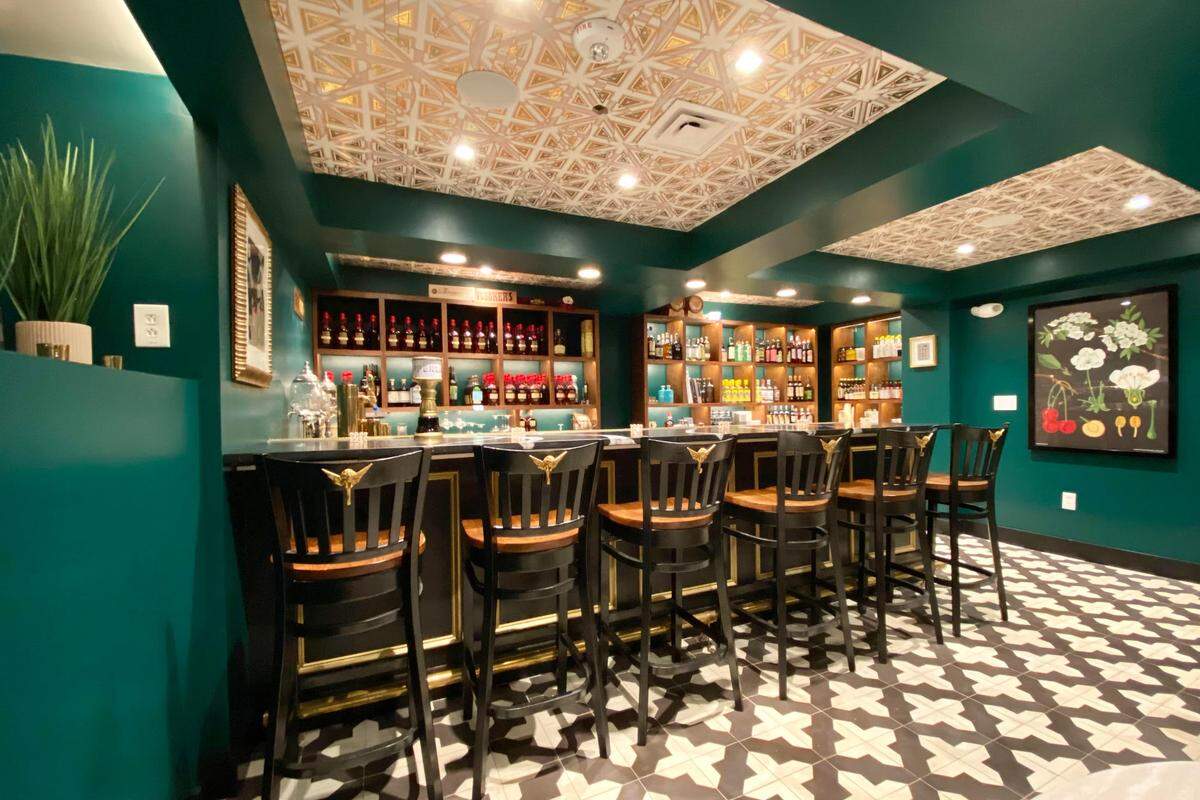 A wide shot of a sophisticated small bar. The walls are a deep teal, the floor is a striking black and white geometric tile, and the coffered ceiling features gold-patterned tin tiles. The long black bar is attended by a row of black bar stools, each with a golden eagle emblem on its back.