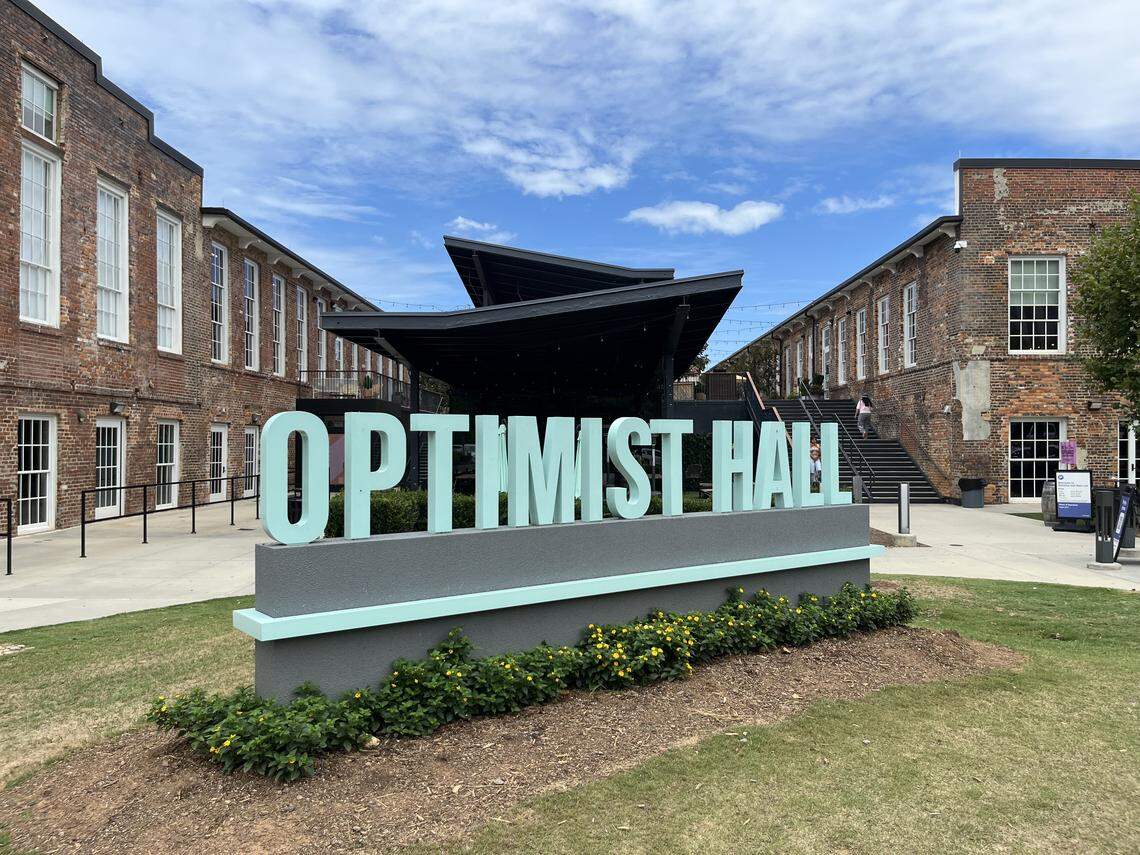 A large, teal-colored outdoor sign reads “OPTIMIST HALL” in bold capital letters. The sign is positioned on a grassy area in front of a courtyard flanked by two historic-style brick buildings.