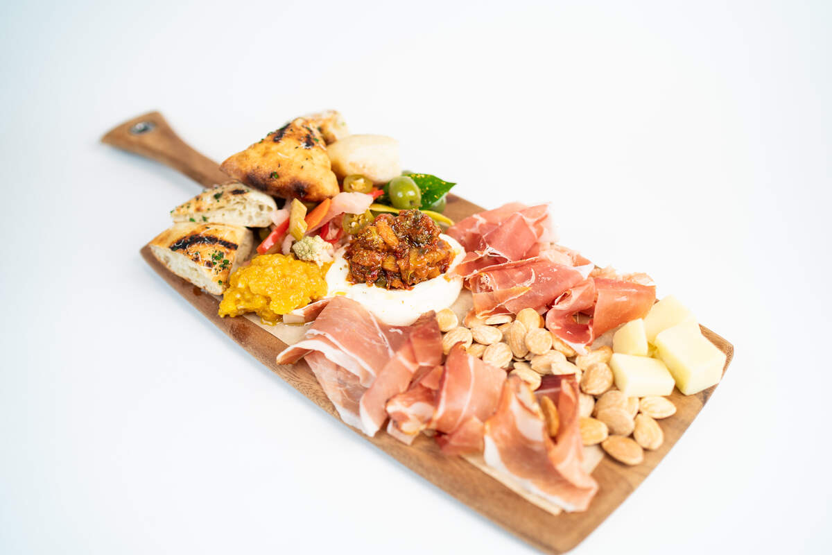 A large wooden cutting board, with a handle, holds a vibrant and abundant charcuterie platter. The board is arranged with thin slices of prosciutto, a pile of whole almonds, and chunks of cheese. In the center, a large dollop of white burrata cheese is topped with a relish or chunky sauce. Grilled bread slices, green olives, pickled vegetables, and a yellow spread, likely a puree or dip, are also artfully arranged on the board. The platter is shot from a slightly elevated angle against a clean white background.