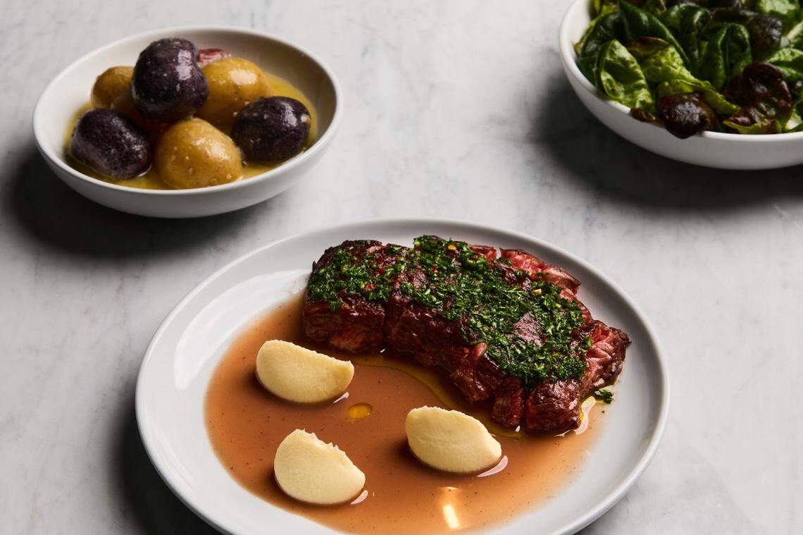A top-down view of a gourmet meal served on a white marble surface. In the foreground, a rectangular white plate holds a perfectly cooked cut of steak, richly red and topped with a generous layer of finely chopped green herbs and what appear to be red chili flakes. The steak rests in a pool of light brown au jus. Three half-moon shaped portions of what looks like potato puree or dumplings are arranged on the plate around the steak. In the upper left, a white bowl contains a mix of whole purple and yellow potatoes. In the upper right, another white bowl is filled with a green leafy salad, possibly mixed greens with red accents.