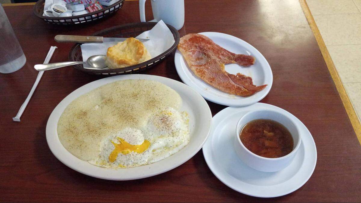A high-angle, close-up shot of a hearty breakfast spread on a dark wood-grain table. The meal includes a large portion of white grits heavily seasoned with black pepper, served alongside two fried eggs with runny yolks. A separate plate holds a large slice of pan-fried country ham. To the bottom right, a small white bowl contains a dark brown liquid, likely red-eye gravy or syrup. A small bread basket contains a single golden-brown biscuit. In the background, there is a plastic basket of condiment packets, a white mug, and a tall glass of water with a white straw.