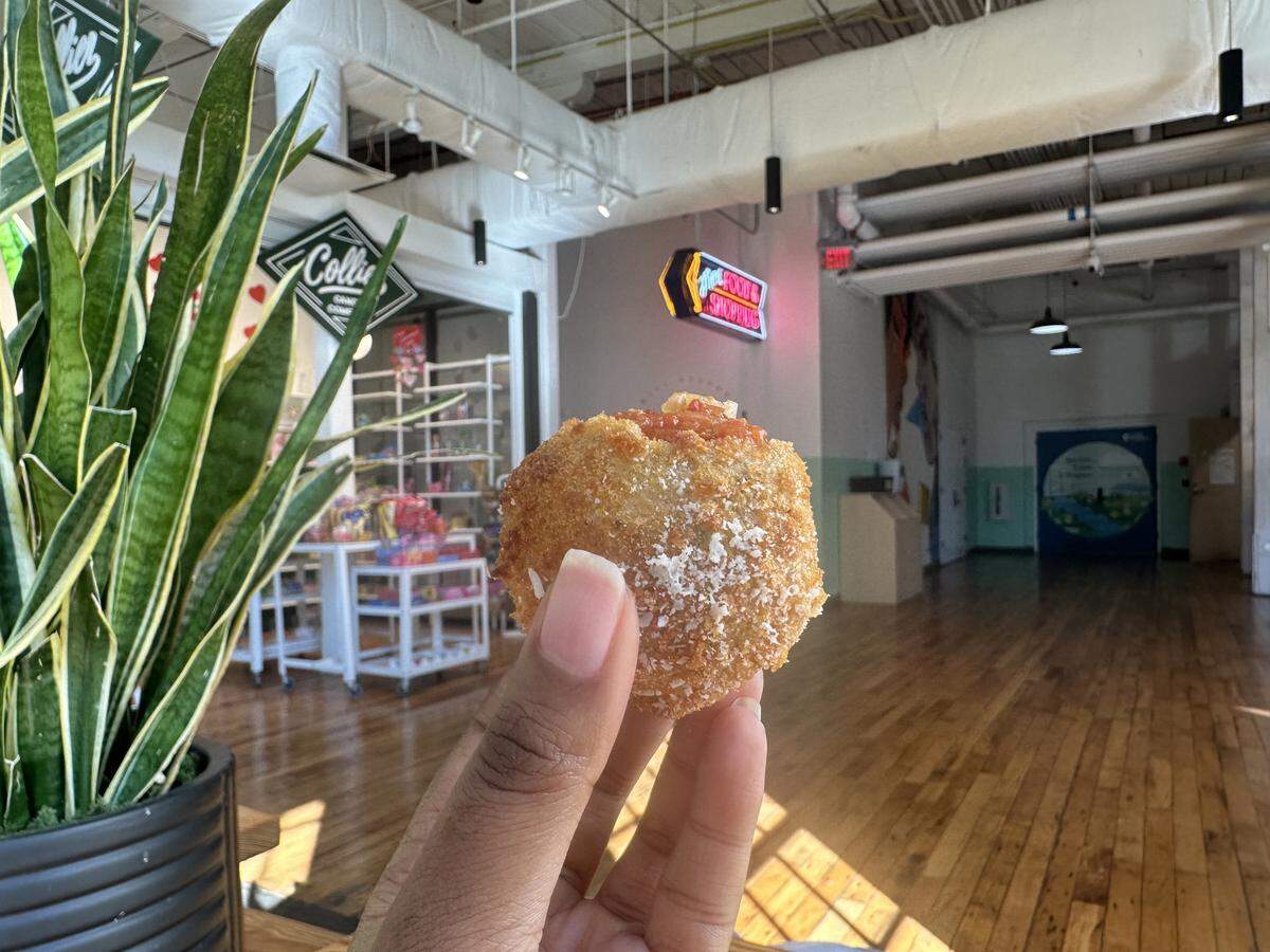 A close-up shot of a hand holding a single, golden-brown fried rice ball (arancini) dusted with white cheese. The background shows a bright, modern indoor market space with polished wooden floors, white industrial ceilings, and a large indoor plant to the left.