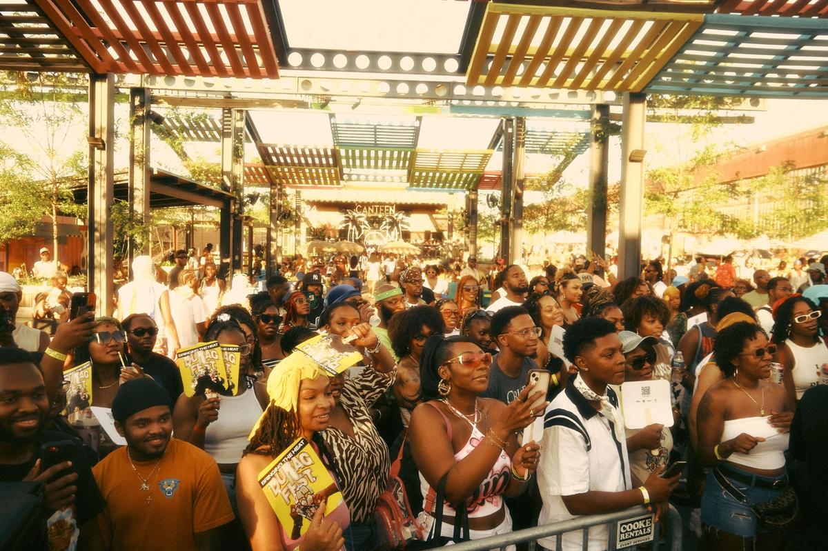 An eye-level, medium shot captures a vibrant, high-energy crowd of mostly Black people at a music festival or outdoor event called “DURAG FEST.” The photo has a warm, vintage aesthetic with golden, film-like tones and a slight grain. In the foreground, a dense group of attendees stands behind a metal barricade. Several people are holding up yellow fans that feature the event’s name and a cartoon graphic.