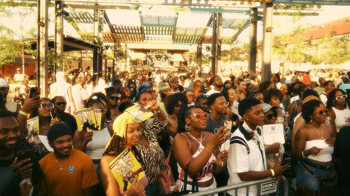 An eye-level, medium shot captures a vibrant, high-energy crowd of mostly Black people at a music festival or outdoor event called “DURAG FEST.” The photo has a warm, vintage aesthetic with golden, film-like tones and a slight grain. In the foreground, a dense group of attendees stands behind a metal barricade. Several people are holding up yellow fans that feature the event’s name and a cartoon graphic.