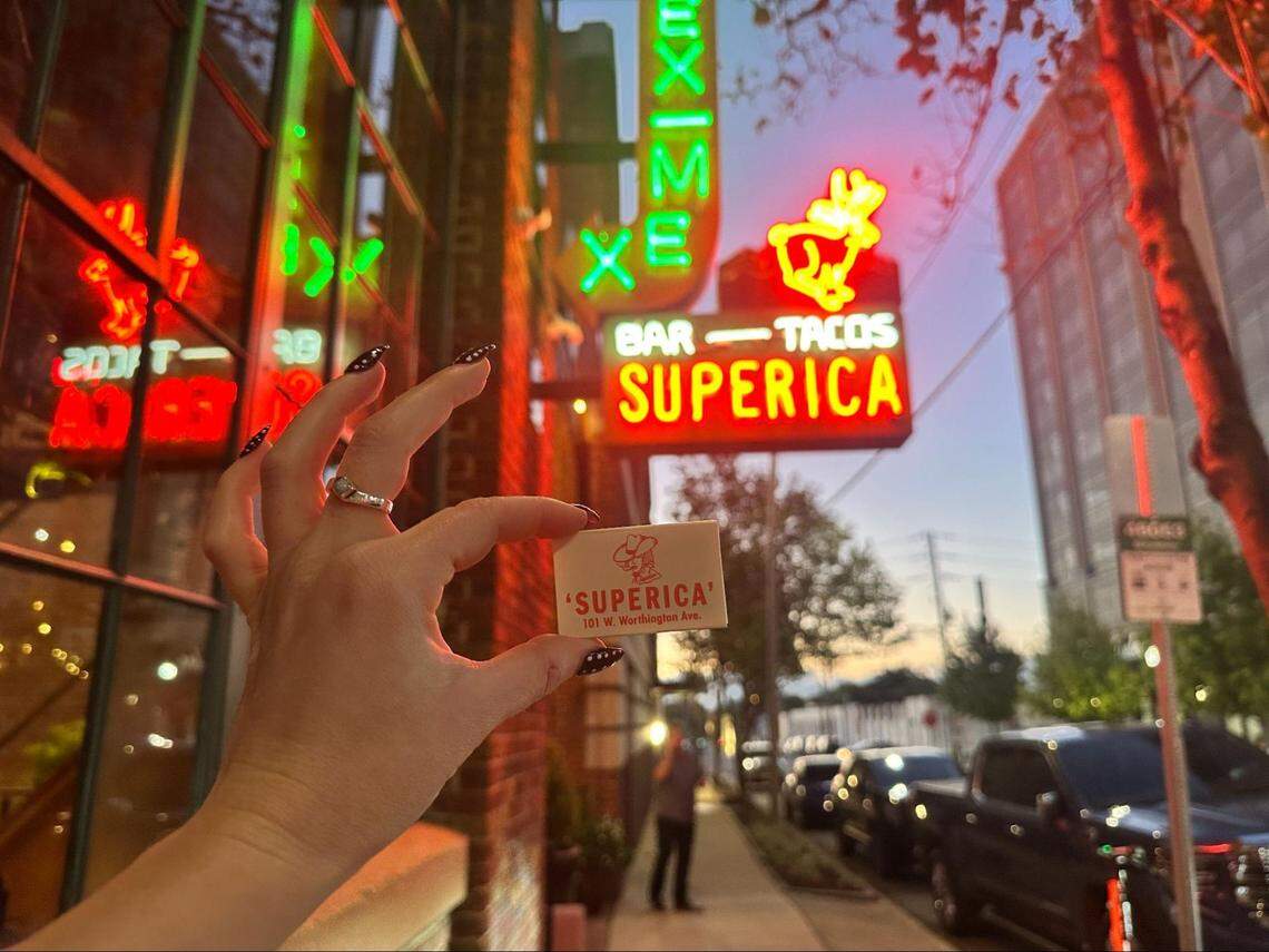 A person’s hand with dark, patterned nail art holds up a white matchbook for “’SUPERICA’” on a city street at dusk. The background is brightly lit by the restaurant’s red and green neon signs, which feature the name “SUPERICA,” “BAR — TACOS,” and a bucking bronco. The warm light from the signs reflects on the brick building and the street scene behind.