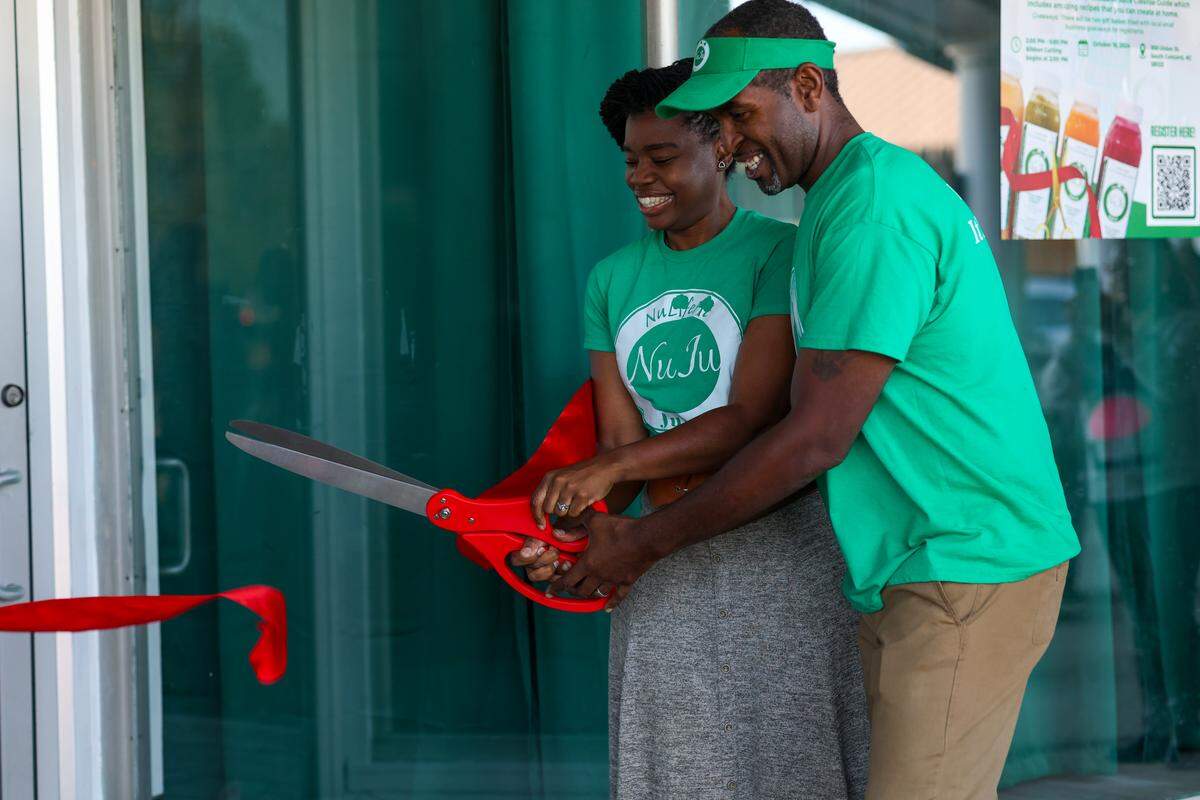A couple, both smiling and wearing matching green “NuJu Juice” t-shirts, stand together in front of a glass storefront. They are jointly holding a pair of giant red scissors, in the process of cutting a thick red ribbon for a grand opening ceremony.