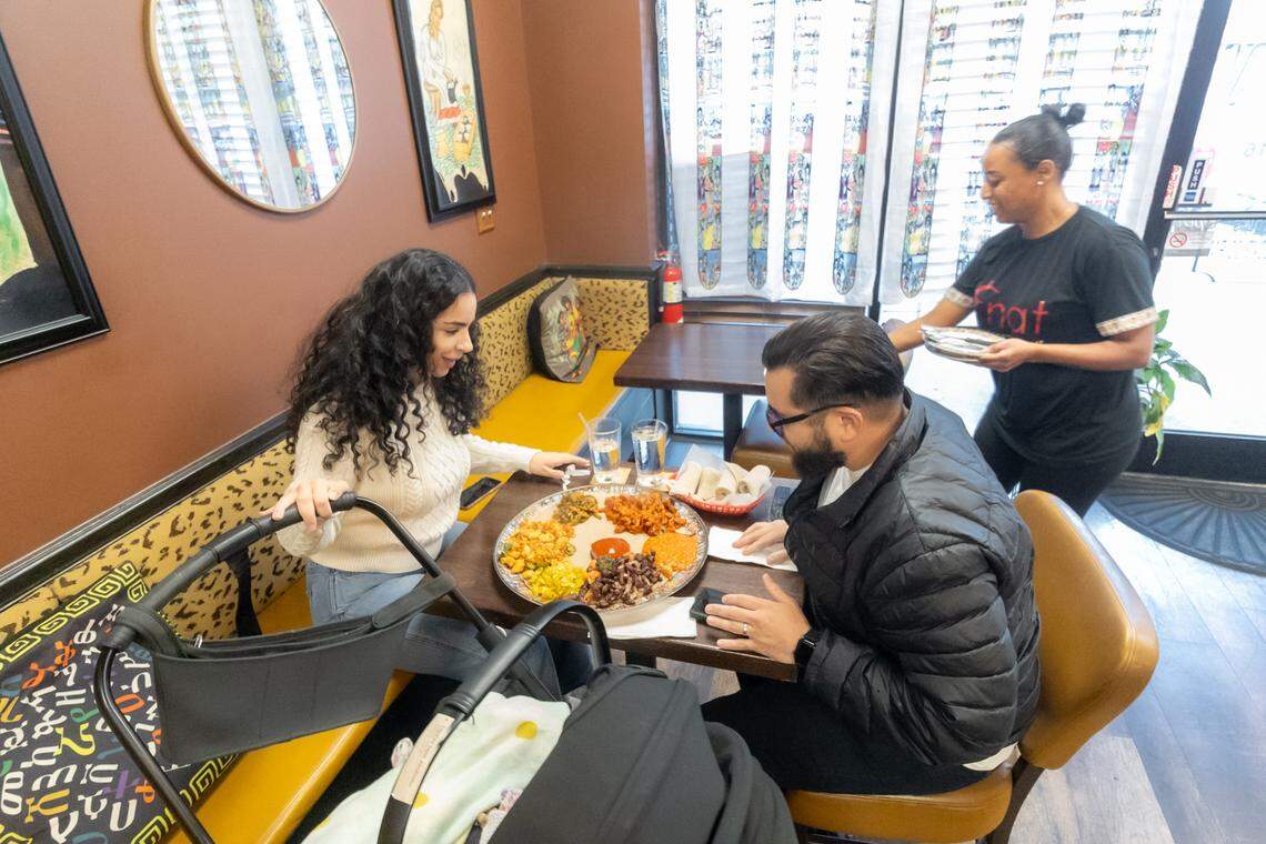 Inside the dining room, a server delivers a fully loaded communal platter to a couple seated at a dark wood booth. The platter is vibrant with various portions of Wot (stew) and vegetables arranged on injera. The dining area features warm brown walls, yellow cushioned seating with patterned accents, and a baby stroller parked by the table, emphasizing a family-friendly environment.