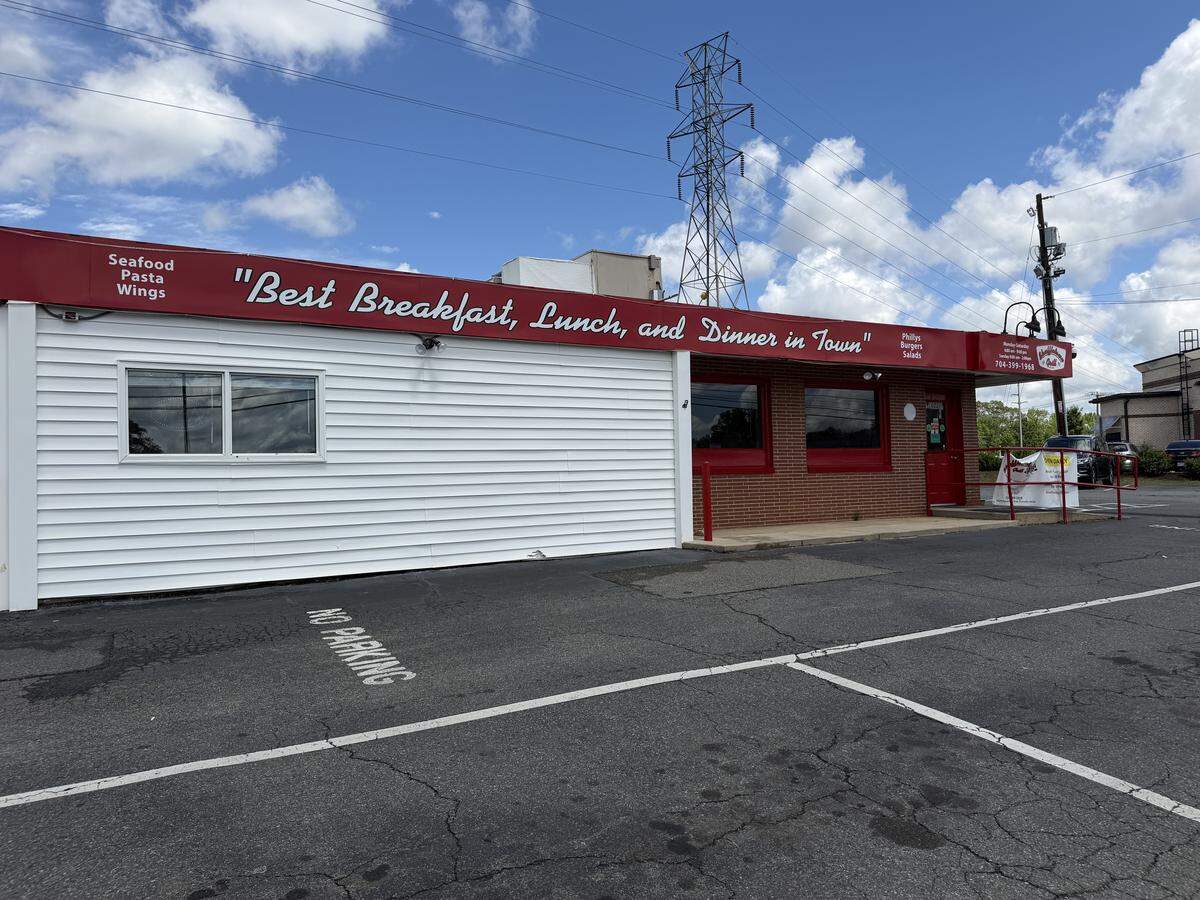 A wide exterior shot of Shuffletown Grill, a long, low-profile building with white siding and a red brick entrance area. A large red awning runs across the top of the building with white script lettering that reads, “Best Breakfast, Lunch, and Dinner in Town.” The parking lot in the foreground is empty, and a tall utility tower stands against a blue sky with white clouds in the background.
