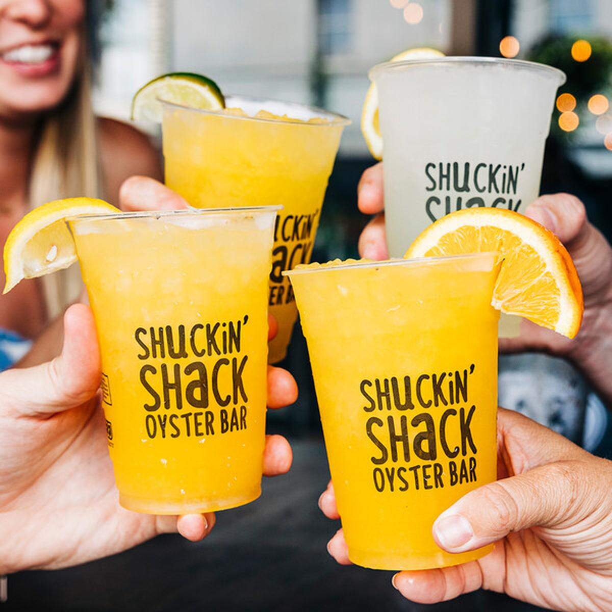 Four people hold up clear plastic cups branded with the “Shuckin’ Shack Oyster Bar” logo in a celebratory toast. Three of the cups are filled with a bright orange citrus drink and garnished with fruit, while one contains a pale yellow beverage.