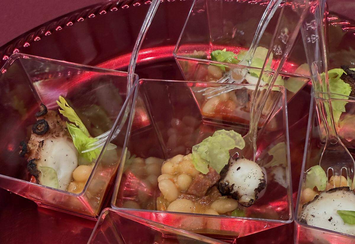 This close-up shot features small appetizer portions of a signature dish from Guard and Grace, likely a preview of their Oak Fired Octopus. Each small, clear plastic square cup contains a single charred piece of octopus tentacle, served alongside white beans, a light tomato-based broth or sauce, and a fresh garnish of celery leaf.