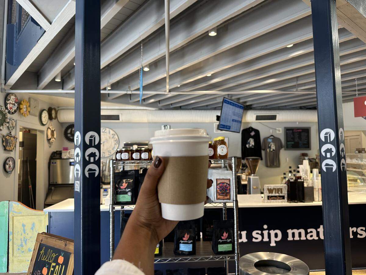 A first-person perspective shot of a hand holding up a white to-go coffee cup with a cardboard sleeve. The cup is in the foreground, and the background is the blurred interior of a coffee shop. The shop features a counter with bags of coffee and jars of honey for sale, a high, beamed ceiling, and a wall decorated with numerous clocks. A small chalkboard sign in the bottom left reads “hello FALL”.
