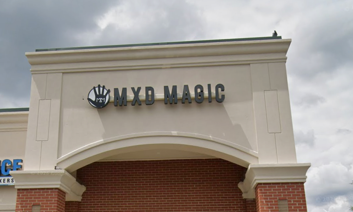 The storefront features a cream-colored architectural archway with a sign that reads “MXD MAGIC” in black, block lettering. Next to the text is a circular logo containing a hand silhouette with “MXM” written across the palm.