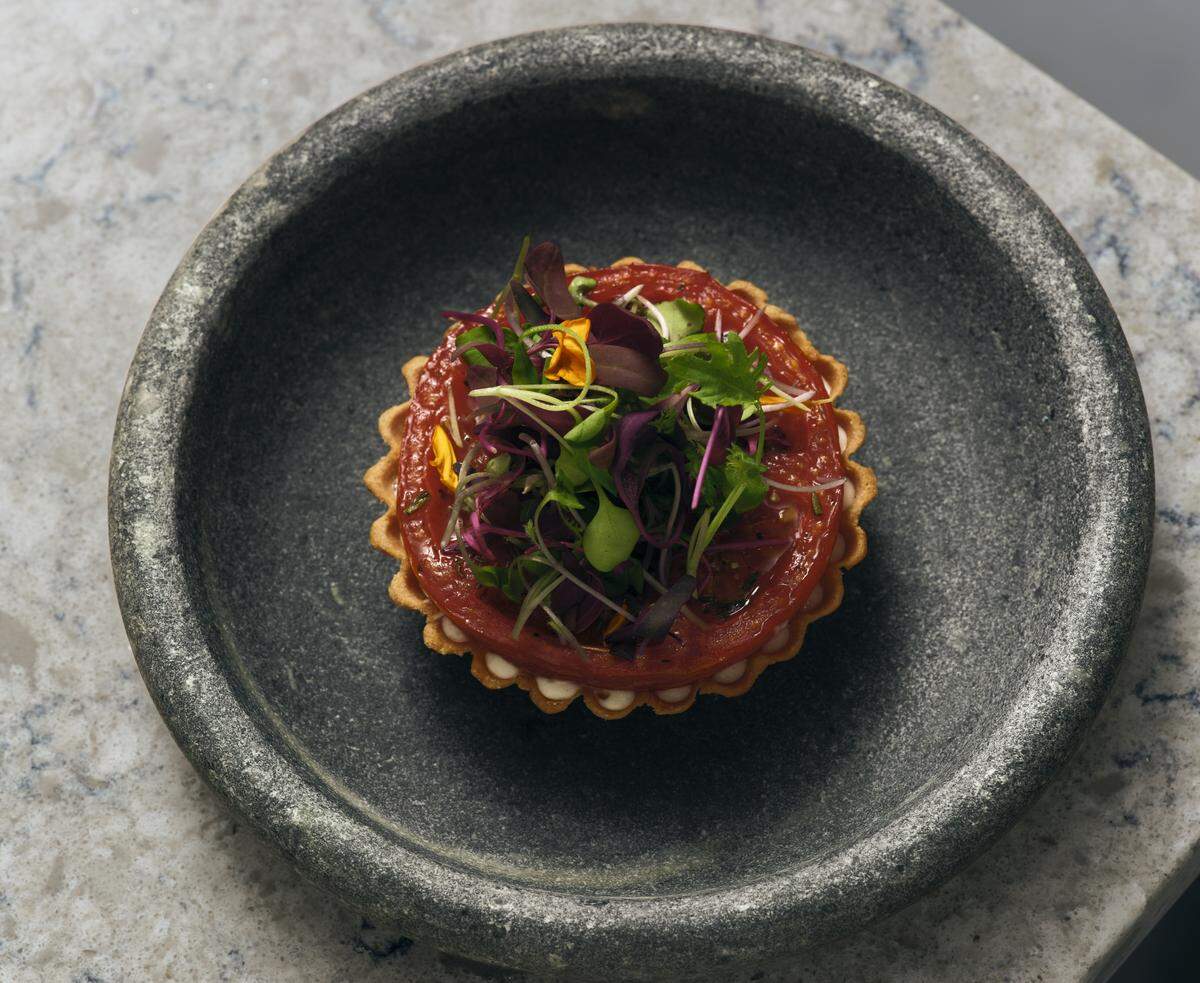A top-down, close-up shot of a gourmet tomato tartlet, artfully presented as a fine-dining dish. The tartlet features a golden, fluted pastry crust filled with a layer of piped white cream dots along the inner edge. It is topped with a thick, vibrant red heirloom tomato slice that acts as a base for a delicate mound of fresh microgreens, including purple and green sprouts and tiny yellow edible flower petals.