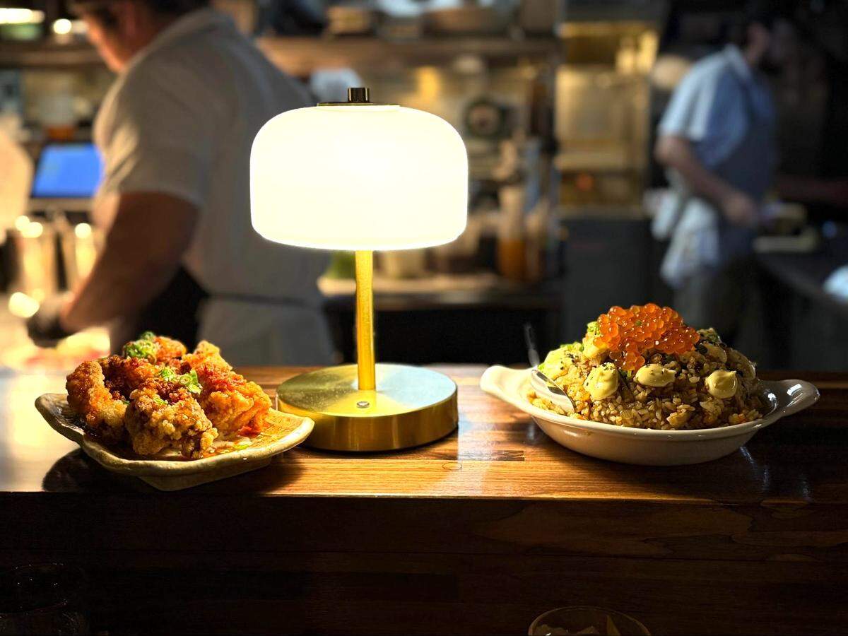 A wide shot of the restaurant’s wooden counter featuring the crispy fried chicken on the left and the seafood fried rice with salmon roe on the right. A warm gold table lamp sits between the plates, with the blurred figures of chefs working in the background.